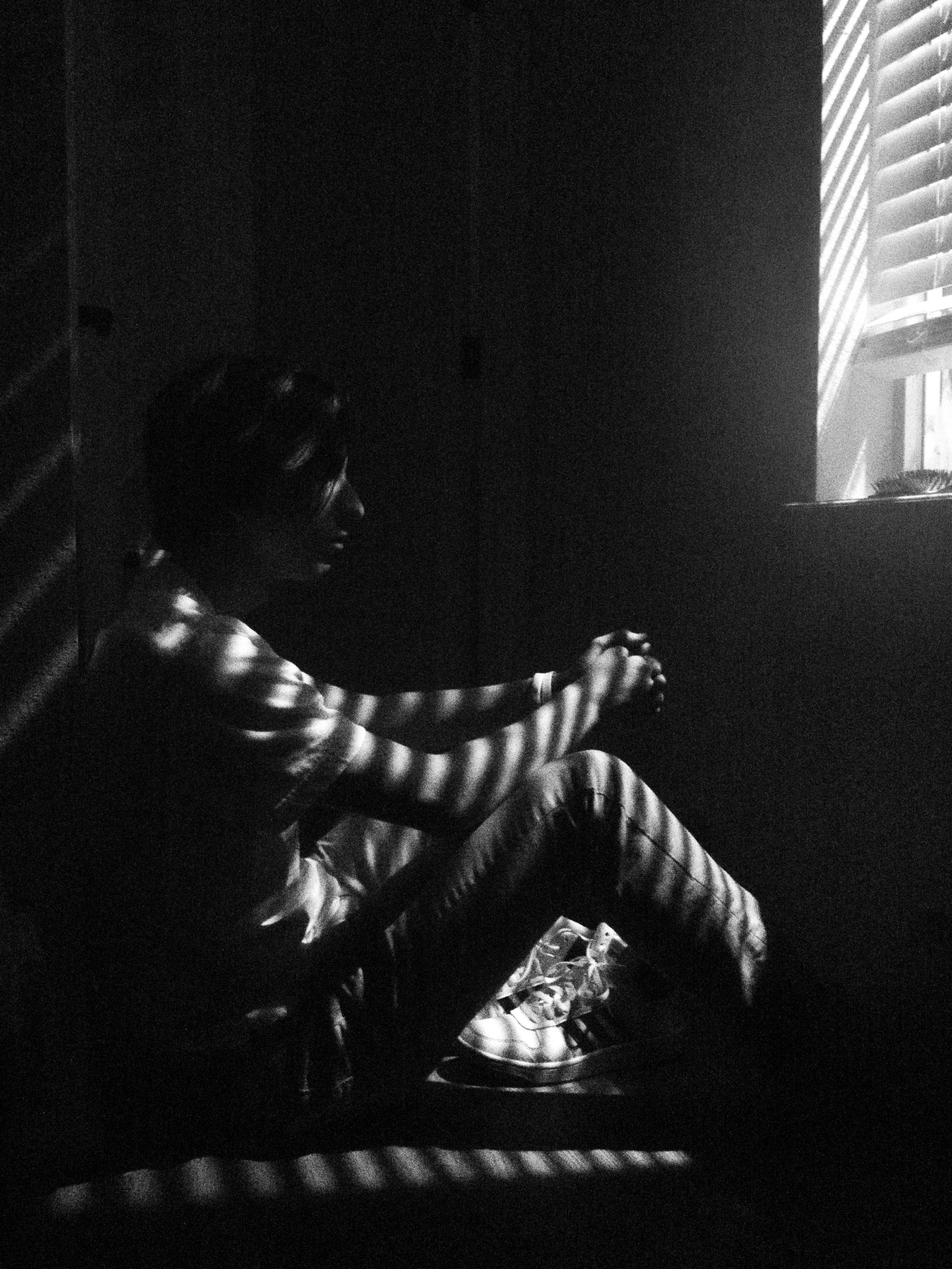 Black and white picture. Man, sad, sitting in front of window, with shutters down, small rays of light shining on him.
